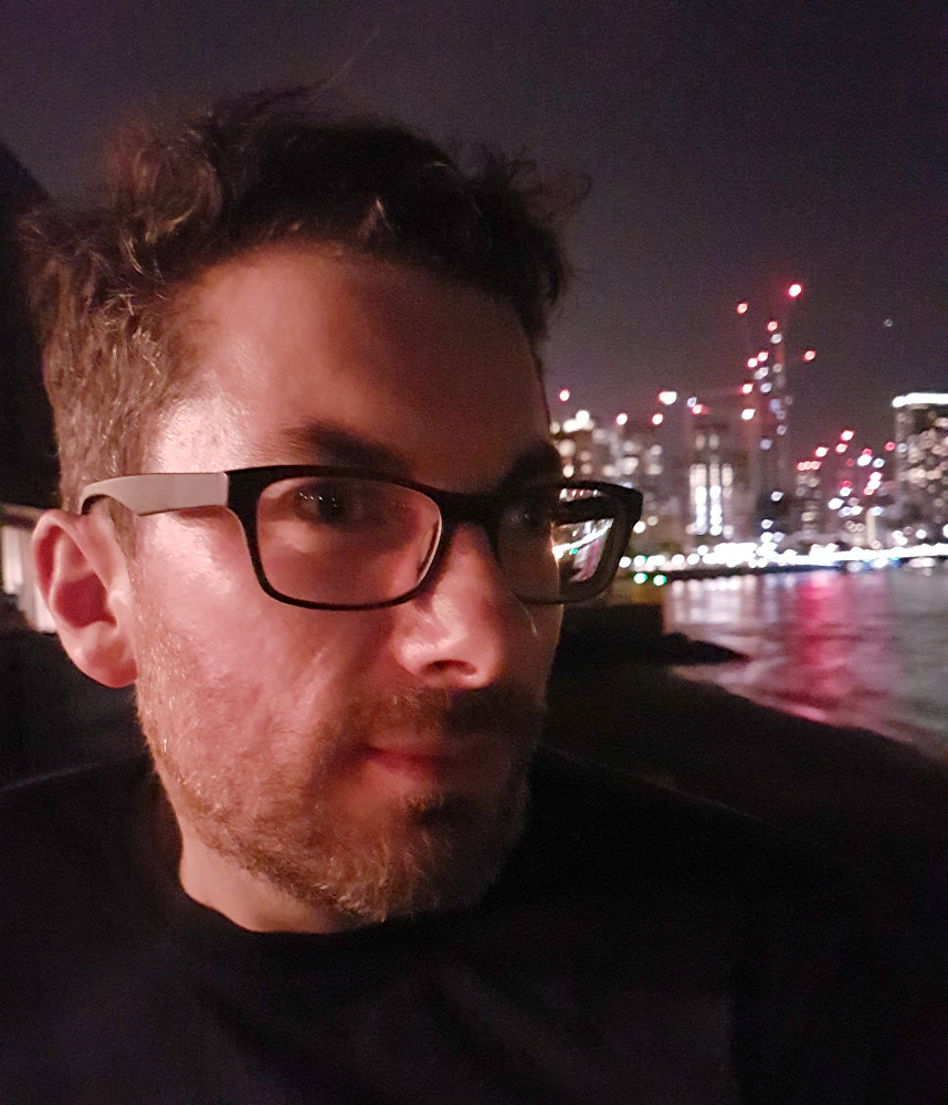 A picture of Matt Webster in a city at night. He has brown hair and glasses.